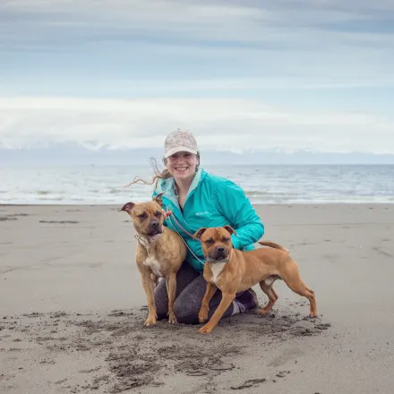 Elora's staff photo from Alaska Veterinary Clinic where she is at a beach with her two brown dogs smiling.